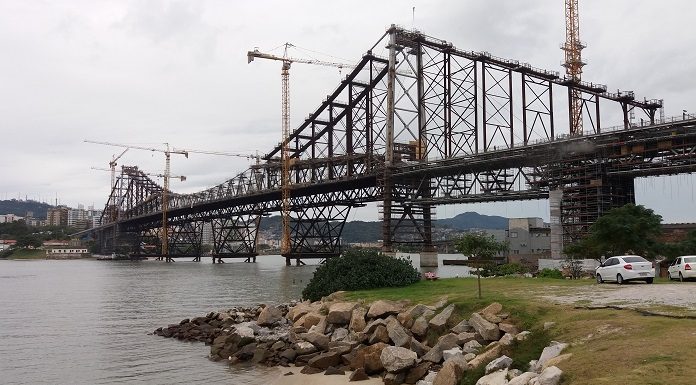 Grande estrutura metálica vista de lado que atravessa um canal, com gruas ao lado da ponte