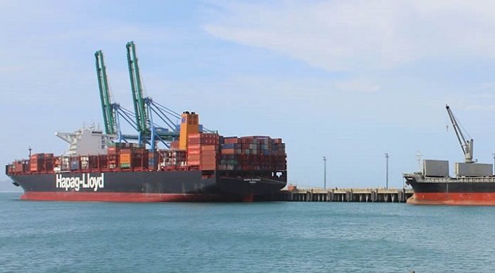 navio cargueiro visto ao longe com containeres atracado em um porto em um dia de sol e mar calmo