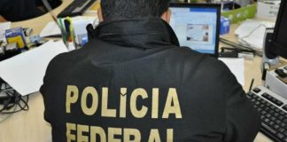 agente visto de costas sentado em uma bancada com laptop usando colete escrito polícia federal