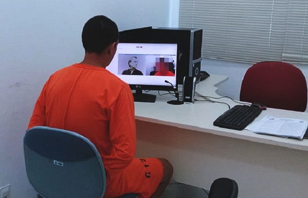 homem utilizando macacão laranja de detento é visto de costas sentado em frente a um computador com imagens de webcams de outras pessoas