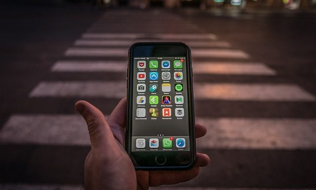 close em uma mão que segura um celular com tela ligada mostrando aplicativos; pessoa está em frente a uma faixa de pedestre