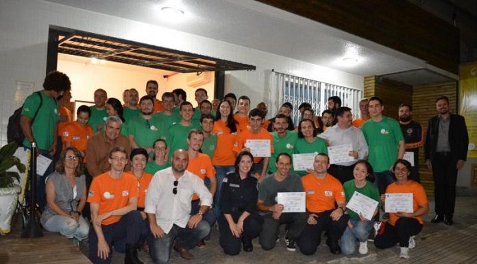 grupo de voluntários, com camisas laranjas ou verdes, posa para a foto de noite. ao centro do grupo e agachada está a secretária de segurança de florianópolis