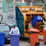 trabalhadores da comcap mexem em latões de lixo atrás de um caminhão de lixo, parado no meio de uma rua