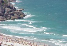 foto de uma praia em dia ensolarado com muitos banhistas vista de cima e de longe, como que de um prédio