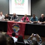 diversas pessoas em uma bancada à frente de um auditório; há uma bandeira do movimento de população de rua sobre a bancada; deputada carminatti ao centro fala no microfone