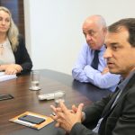 moisés, daniela e dado cherem sentado na ponta de uma mesa; carlos moisés está falando e gesticulando