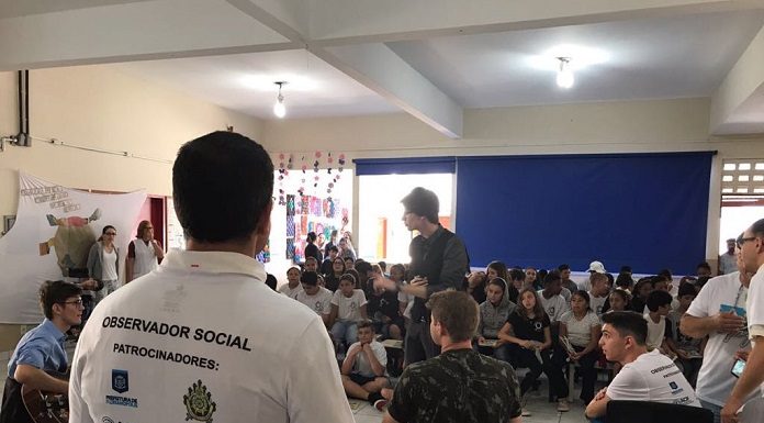 homem visto de costas com camisa escrita "observador social" fala para um grupo de estudantes