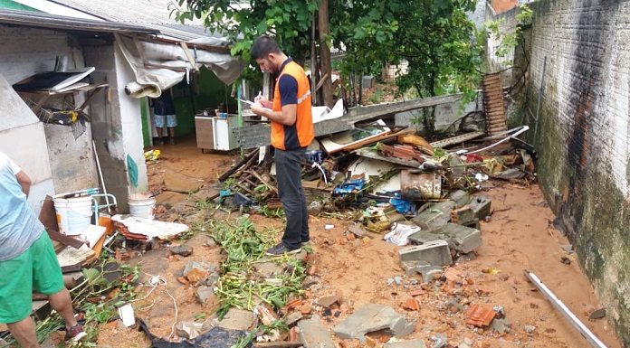 funcionário da defesa civil usando o colete laranja anota em prancheta enquanto em pé em área de lama ao lado de casas atingidas