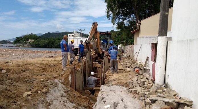 área de areia com vala aberta por escavadeira e operarios de capacete trabalhando