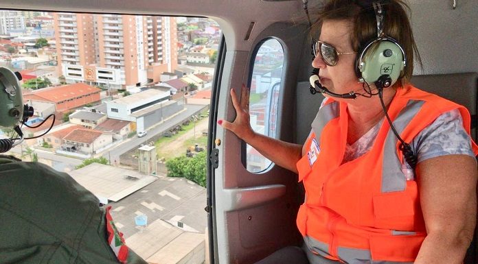 adeliana usando colete, óculos escuros e fone ouvido dentro do helicóptero ao lado da porta aberta olhando para fora, com a cidade em baixo; um militar ao lado