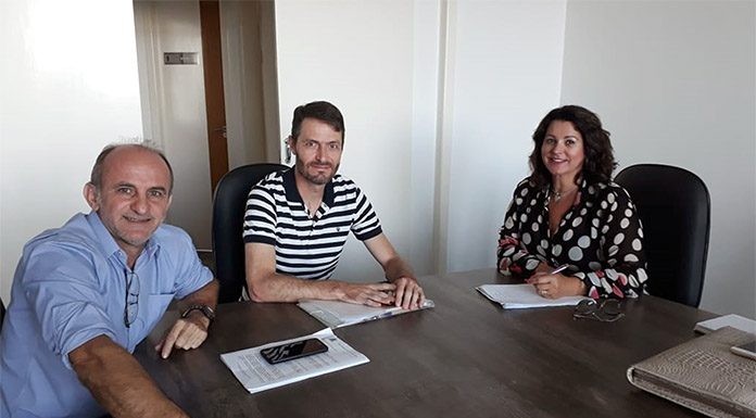 os dois vereadores e a vereadora sentados na ponta de uma mesa em uma sala olham para a câmera