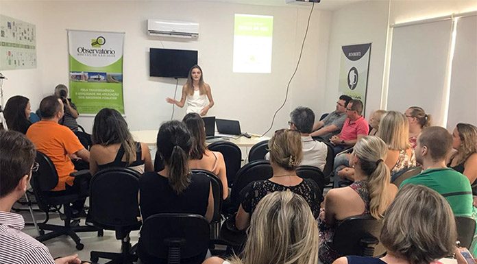 em uma sala lotada de pessoas sentadas, mulher dá palestra ao lado de um banner do ossj