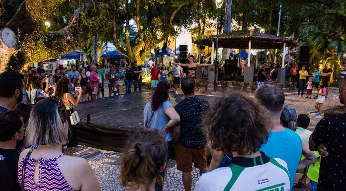 público de costas para a câmera assiste um show no coreto da praça, debaixo de muitas árvores