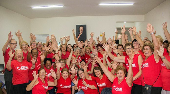 grande grupo de voluntárias usando a camiseta vermelha da associação posa de braços para o alto para a foto; a maioria idosas