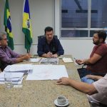 prefeito na ponta de uma mesa de mármore conversa com as demais pessoas em uma sala que tem as bandeiras do brasil, santa catarina e biguaçu