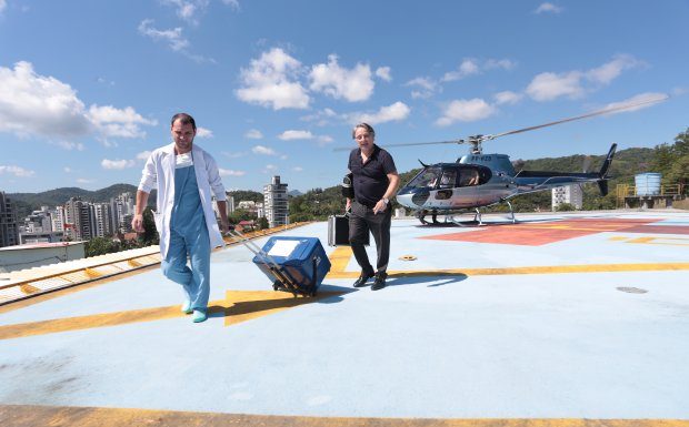 médico carrega caixa térmica por um heliponto no alto de um prédio em balneário camboriú; helicóptero ao fundo e outro homem