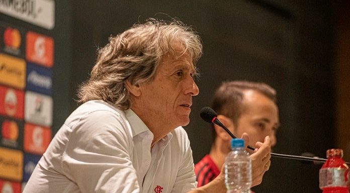 jorge jesus fala ao microfone gesticulando; há uma garrafa de água na bancada e uma pessoa atrás