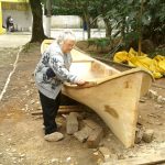 a senhora de pantufas lixa a borda da canoa parada sobre troncos em uma área de terra com árvores ao fundo