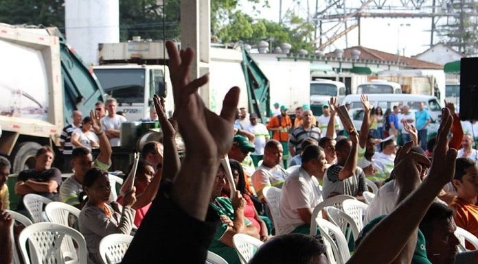 trabalhadores da comcap em cadeiras com as mãos levantadas em votação e caminhões de lixo ao fundo