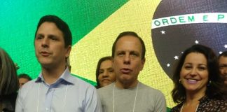 três pessoas lado a lado com a bandeira do brasil em telão ao fundo