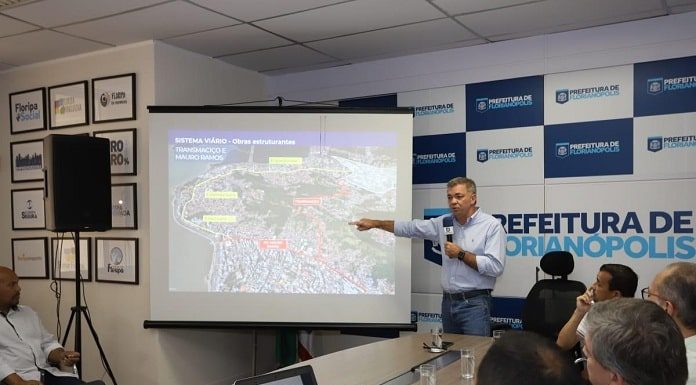 gean discursa em pé com microfone na mão apontando para uma imagem projetada de mapa da cidade