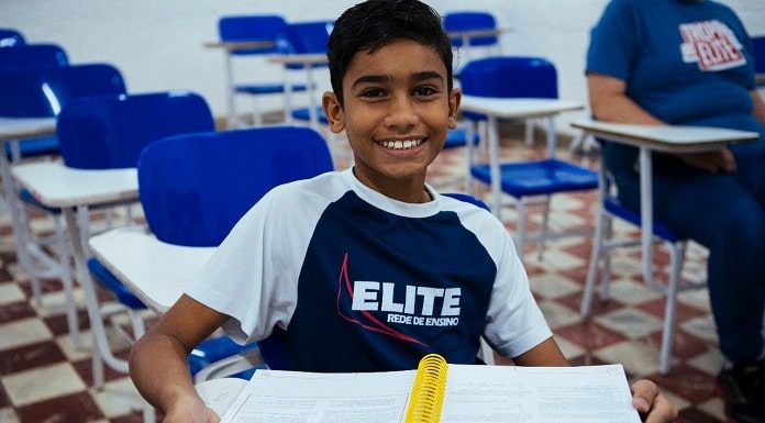 aluno, sentado em carteira de sala de aula vazia, sorri para a câmera com uniforme do elite ceb