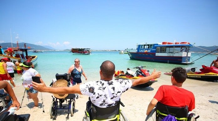 homem em cadeira de rodas com os braços abertos na beira da praia da ilha do campeche e outras pessoas, em pé ou cadeiras de rodas, em volta; barcos na água