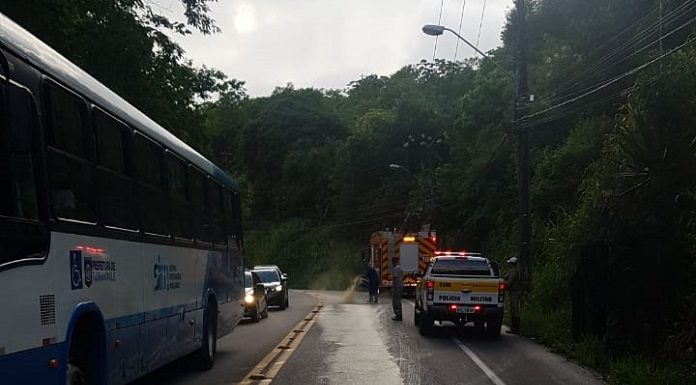 bombeiro limpando o asfalto com mangueira e marca de óleo que desce a via, ao lado de ônibus, caminhão dos bombeiros e viatura da polícia