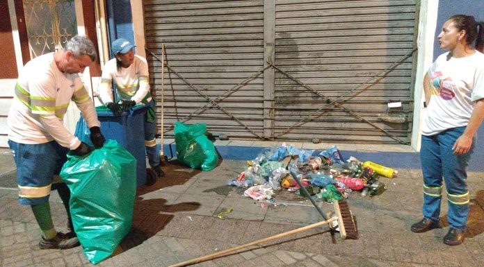 um funcionário e duas funcionárias da comcap recolhem lixo de um calçadão em frente a comércio; homem fecha sacola de lixo