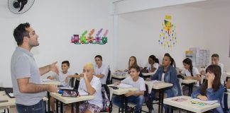 professor fala e gesticula em pé na frente da sala com alunos e alunas sentados nas carteiras