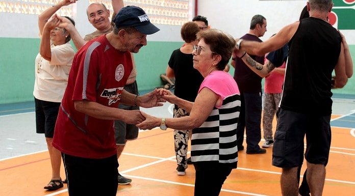 homem e senhora dançam de mãos dadas em quadra de esportes com outras pessoas dançando em volta