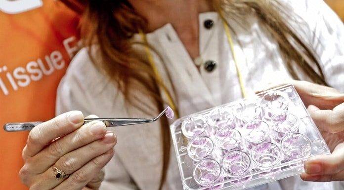 mulher segura com uma pinça um pedaço do produto de frente para a câmera e na outra mostra placa, similar a forma de gelo, com demais produtos