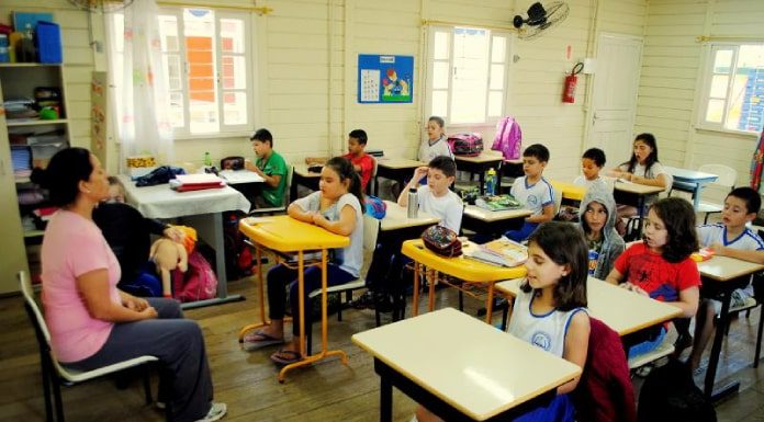 crianças sentadas em carteiras em sala de aula de madeira com professora à frente