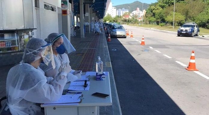 dois profissionais de saúde sentados em mesinha na plataforma do terminal à espera de carros; carros ao fundo; cones, viatura da guarda ao lado