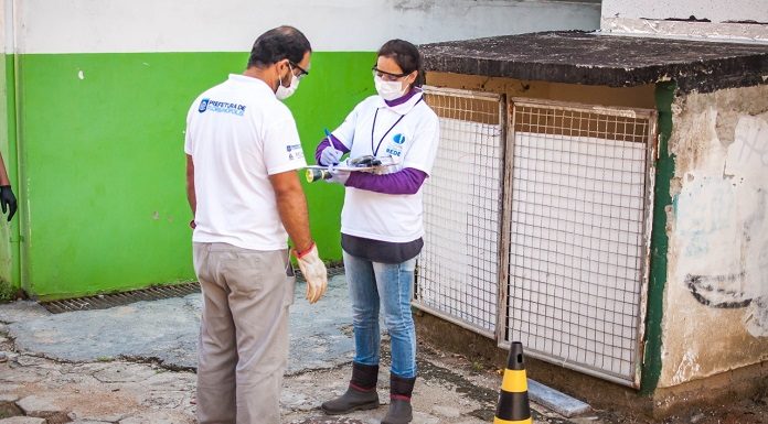 homem e mulher usando equipamentos de proteção e uniformes do programa em pé ao lado de bueiro aberto, com cone, e a mulher aponta algo na prancheta; estão atrás de uma construção