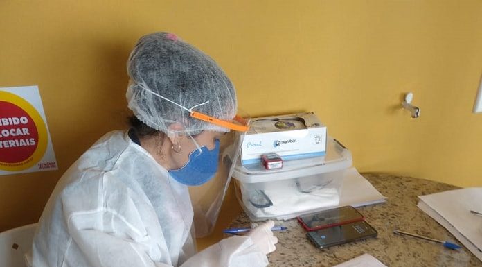 mulher vestida de branco, usando máscara de proteção sentada em uma cadeira escrevendo em papéis em cima de uma mesa