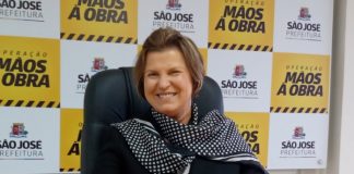 adeliana sorridente com as mãos sobre uma mesa; ao fundo painel com logos da prefeitura de são josé