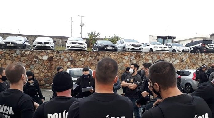 diversos policiais civis vistos de costas reunidos em pé em estacionado; nome da pc nas costas