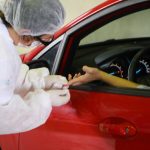 profissional de saúde coleta amostra de sangue de dedo de mulher com mão para fora de um carro vermelho