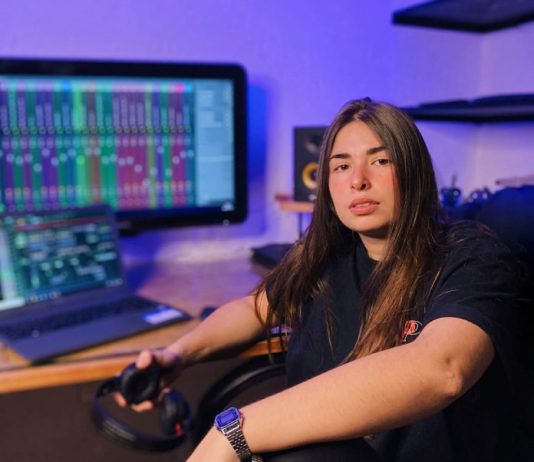 dj katrip posa para a foto em seu escritório, em frente a uma grande tela de computador; ela está com um braço sobre uma das pernas em cima da cadeira