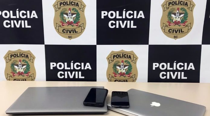 dois notebooks e dois celulares sobre mesa com painel da polícia civil ao fundo