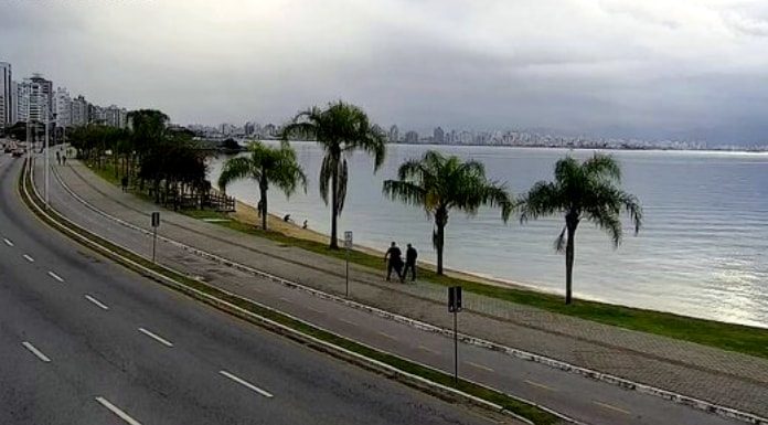 dois homens são vistos andando na beira-mar de florianópolis em foto à distância; palmeiras na orla, mar calmo, sem carros na avenida