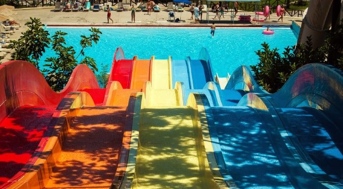 tobogãs coloridos em queda para uma piscina ao fundo, com banhistas circulando; sombra de árvore por cima do tobogã