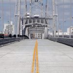 pista da ponte hercílio luz vista de frente com linha divisória de tráfego ao longo da estrutura; base da torre no fundo da foto; prédios ao fundo