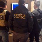 três agentes vistos de costas em pé em sala de apartamento olhando par uma televisão; as inscrições nos casacos de cada um são Dei, Perícia Oficial e Oficial IGP