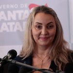 daniela reinehr em frente a um microfone com painel de logos ao fundo