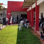 gramado com cadeiras em frente ao cati onde há pessoas em volta usando máscaras esperando em filas e sentadas em volta