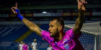 jogador do avaí corre na lateral de campo com os braços levantados comemorando usando camisa rosa; gol e campo com joadores ao fundo; estádio vazio