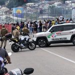 carro jeep da polícia militar atressado na pista com moto lado; motoqueiros em primeiro plano no trânsito. ao fundo no centro da foto grupo protestando e alguns policiais em volta; morro com casas ao fundo