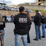 Mecanismo verde: diversos policiais civis homens e mulheres com jaquetas da corporação reunidos em pé em estacionamento; maioria de costas para foto